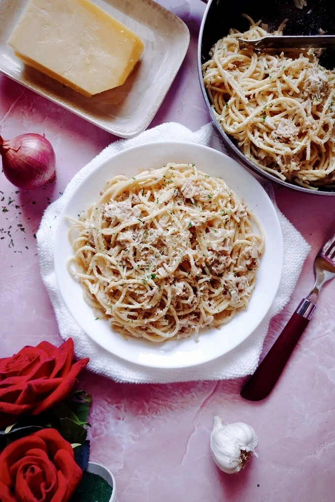 BEST and EASY Creamy Garlic Canned Tuna Pasta Recipe in a white plate and skillet with a whole garlic, an onion, a Parmesan cheese block, a fork and two red roses around it