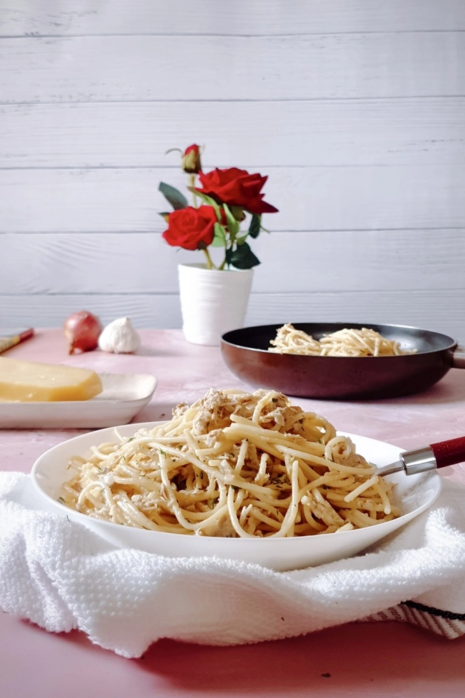 Creamy garlic canned tuna pasta on a white plate over a white cloth in front with a block of Parmesan cheese, whole garlic, whole onion, a white pot of roses and a skillet with tuna pasta in it in the background