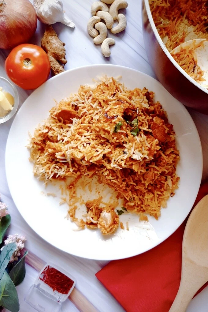 Butteriest Scrumptious Butter Chicken Biryani on a white plate surrounded by ingredients like a tomato, onion, garlic, ginger, cashew nuts and saffron and a wooden ladle on a red cloth