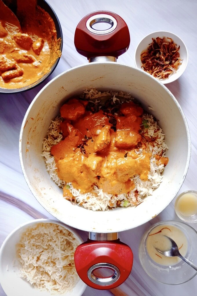 Butter chicken curry spread over biryani rice in a dutch oven surrounded by fried onions, biryani rice, saffron milk, ghee and butter chicken curry, all in separate bowls