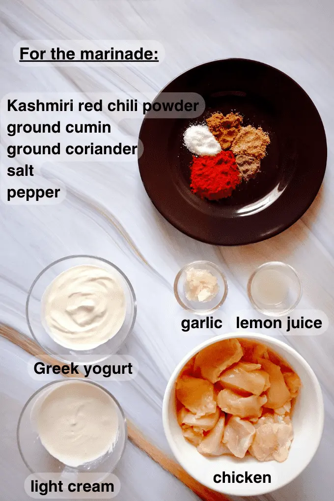 Kashmiri red chili powder, ground cumin, ground coriander, salt and pepper spread on a black plate along with Greek yogurt, light cream, garlic, lemon juice and raw chicken cubes in separate bowls