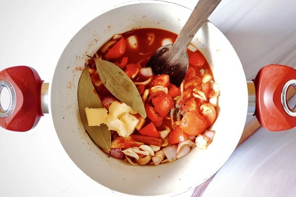 Raw ingredients like chopped onions, cashew nuts, chopped tomatoes, cloves, cinnamon, garlic, ginger, Kashmiri red chili powder, bay leaves, cardamoms, butter, salt and pepper in a Dutch oven with a wooden ladle