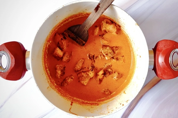 Cooked chicken pieces mixed in with butter chicken sauce in a Dutch oven with a wooden ladle