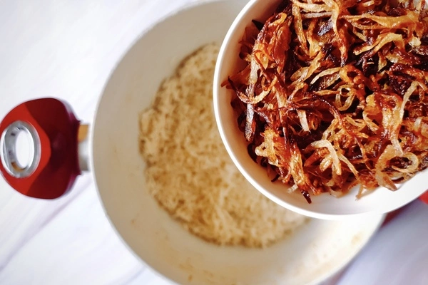 A bowl of fried onions held over a Dutch oven with biryani rice