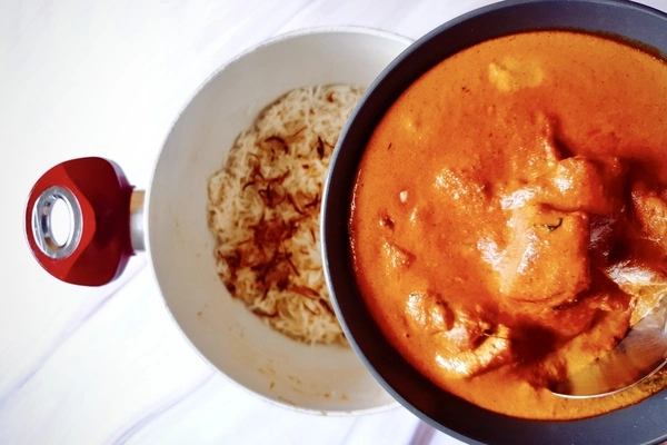 A bowl of butter chicken curry held over a Dutch oven with biryani rice and fried onions in it