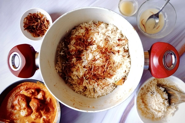Fried onions spread over biryani rice in a Dutch oven surrounded with butter chicken curry, fried onions, ghee, saffron infused milk and biryani rice in separate bowls