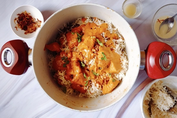 Butter chicken curry and fresh cilantro spread over biryani rice in a Dutch oven surrounded with fried onions, ghee, saffron infused milk and biryani rice in separate bowls