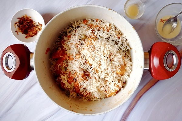 Biryani rice spread over butter chicken biryani in a Dutch oven surrounded with fried onions, ghee, saffron infused milk in separate bowls