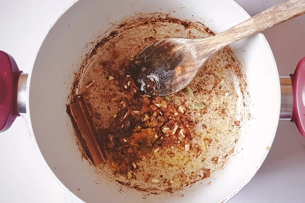 Cinnamon stick, cardamoms, ground cumin and turmeric mixed with onions and garlic in a cooking pot with a wooden ladle