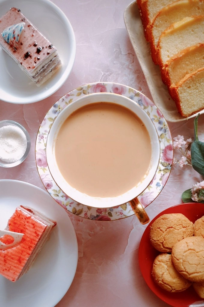 Easy Homemade Japanese Royal Milk Tea Recipe in a tea cup surrounded by cookies, pastries, sliced butter sponge cake, sugar and a flower