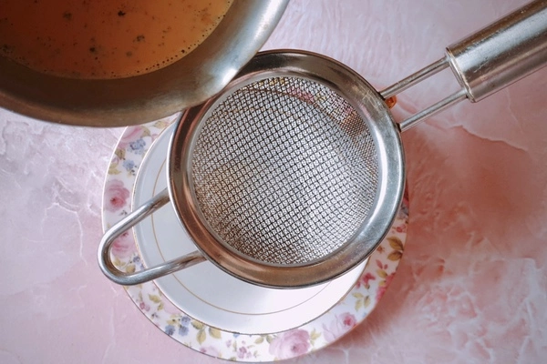 A teacup with a little sugar in it over a saucer with a fine mesh strainer over it