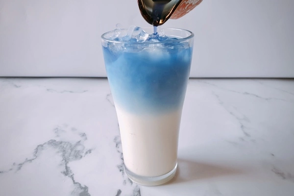 Blue butterfly pea tea being poured into a tall glass with milk mixture and ice cubes in it forming two separate layers of blue tea and milk mixture