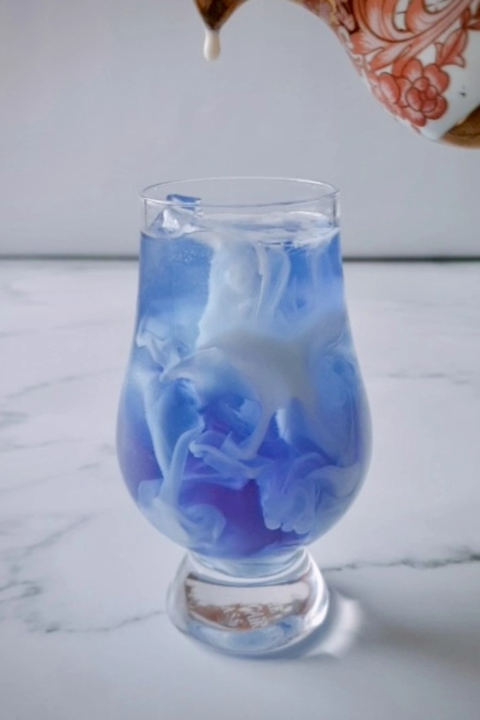 Milk being poured into a glass with blue butterfly pea tea and ice cubes in it showing gorgeous milk swirls