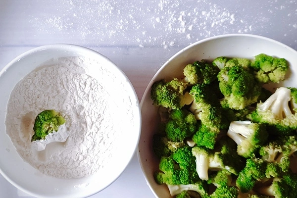 A bowl with the flour mixture and a half coated broccoli floret in it beside another bowl of steamed broccoli florets
