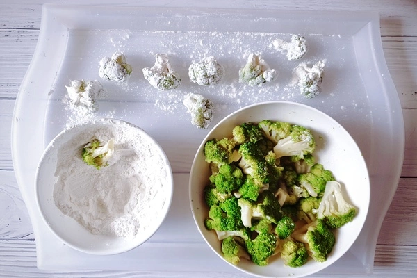 A bowl with the flour mixture and a half coated broccoli floret in it beside another bowl of steamed broccoli florets with some finished coated broccoli florets on a lightly cornstarch dusted tray