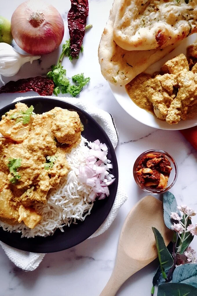 Rich and Aromatic Indian Chicken Korma Curry Cooked in Yogurt on a black plate with rice and chopped onions and on another white plate with two garlic naans also with some vegetables, garlic pickle, wooden ladle and a pink flower on the side