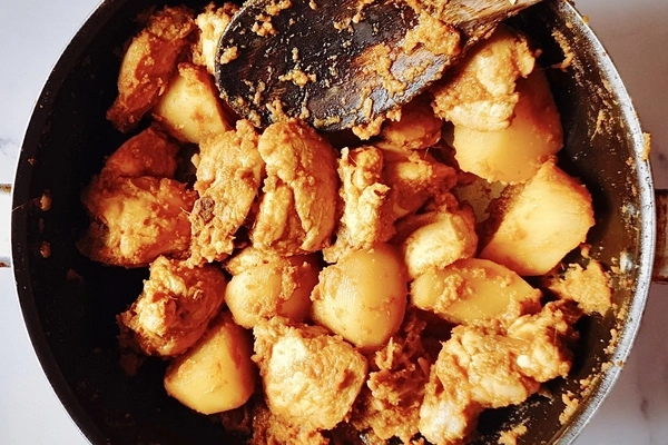 Raw chicken, turmeric, Kashmiri red chili powder, raw potatoes and salt mixed together in a cooking pan with a wooden ladle