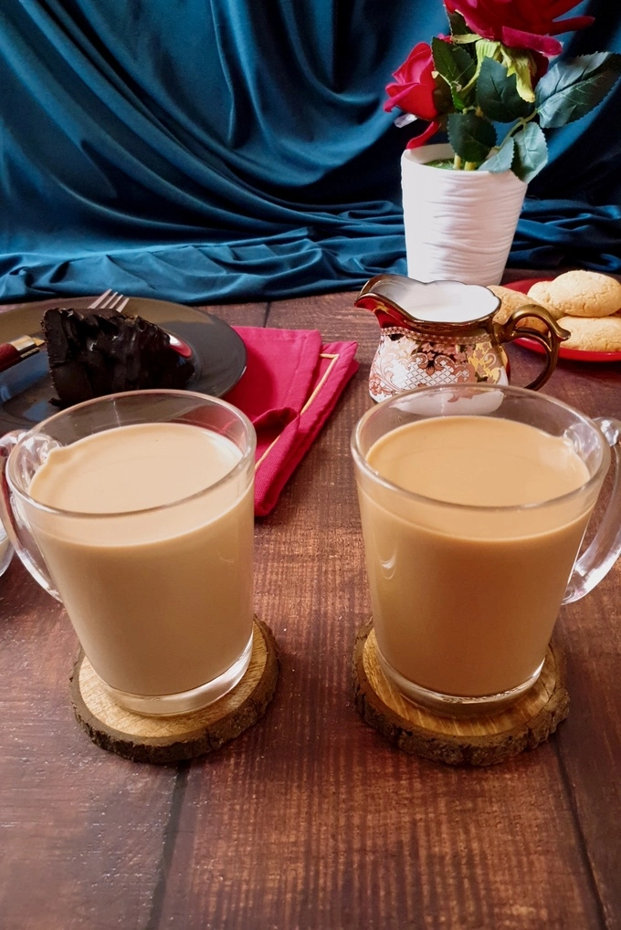 Royal milk tea and regular milk tea in two glass mugs with the background having a black plate with a slice of chocolate cake on it, a milk pourer, a vase of red roses and a small red plate with cookies