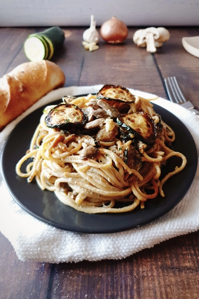 One Pot Zucchini Mushroom Pasta in a black plate placed over a white cloth with bread and a fork on each side and the background having raw ingredients like sliced zucchini, garlic, onion and mushrooms