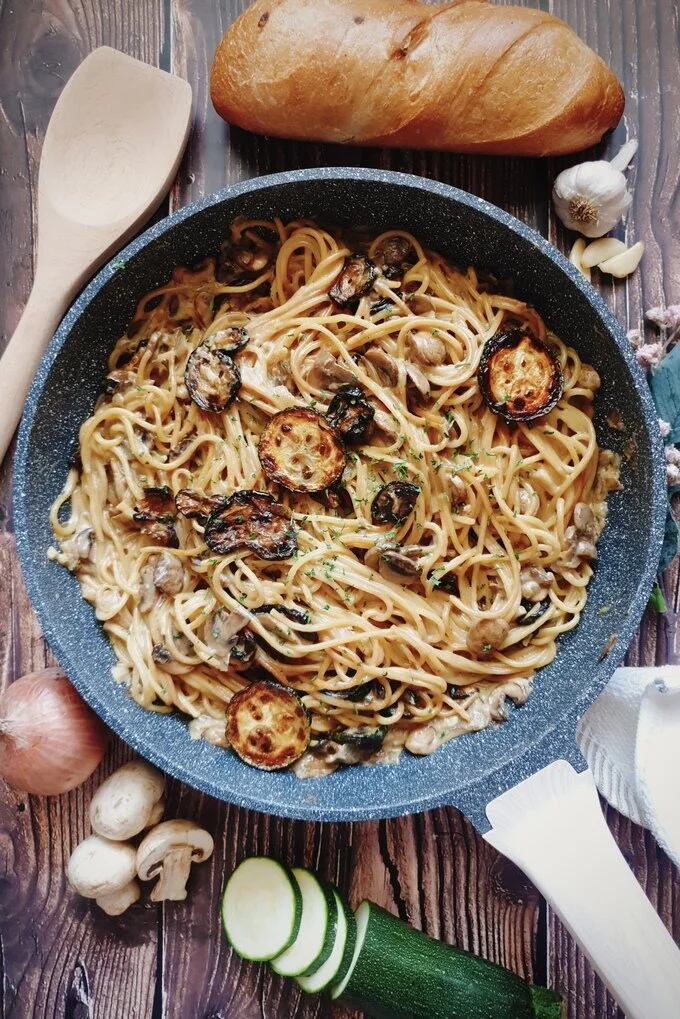 One Pot Zucchini Mushroom Pasta in a skillet surrounded by a wooden ladle, bread, garlic, garlic pods, white cloth, sliced zucchini, mushrooms and an onion