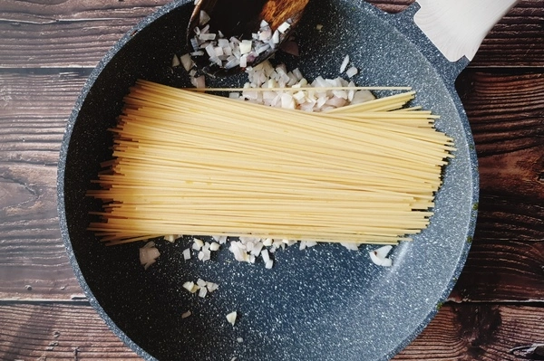 A skillet with uncooked spaghetti, diced onions and chopped garlic in it