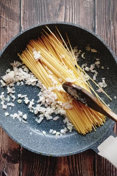 A skillet with diced onions, chopped garlic and uncooked spaghetti being mixed and toasted in it
