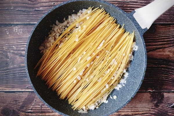 A skillet with diced onions, chopped garlic and uncooked toasted spaghetti spread in it