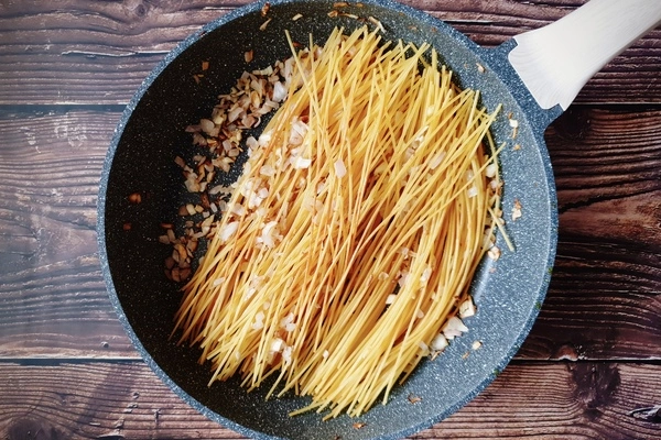 A skillet with diced onions, chopped garlic and browned uncooked toasted spaghetti spread in it