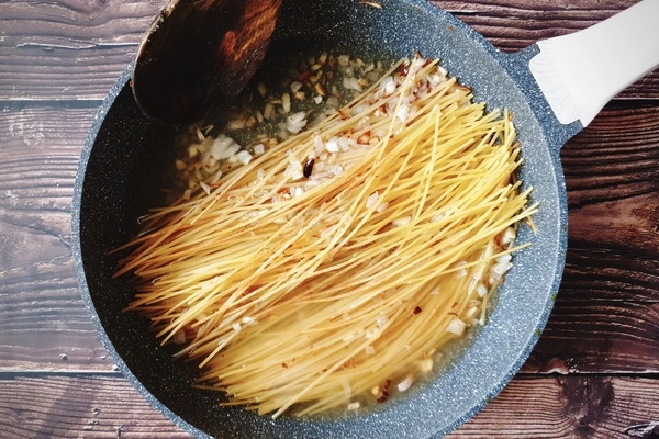 A skillet with diced onions, chopped garlic, browned uncooked toasted spaghetti and vegetable broth in it