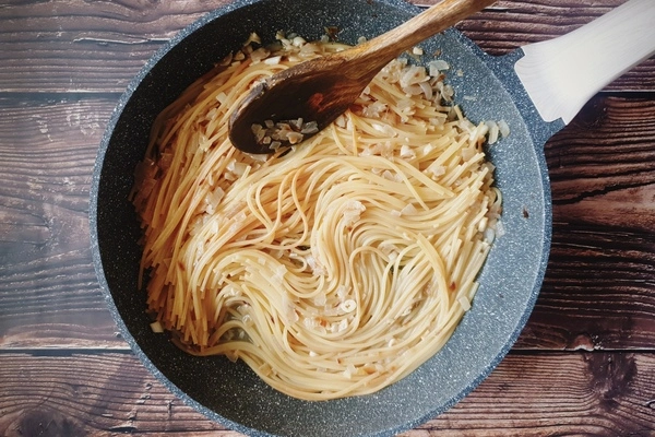 A skillet having diced onions, chopped garlic and cooked spaghetti in it
