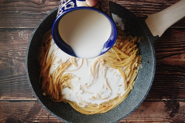A skillet having cooked spaghetti in it with coconut milk being poured in using a blue cup