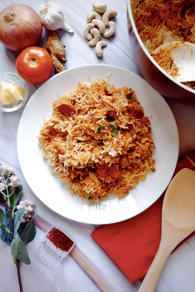 Butteriest Scrumptious Butter Chicken Biryani served on a white plate surrounded by ingredients like a tomato, onion, garlic, ginger, cashew nuts and saffron and a wooden ladle on a red cloth