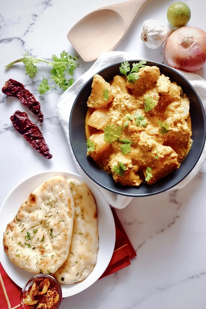 Rich and Aromatic Indian Chicken Korma Curry Cooked in Yogurt served in a black bowl and two naan bread on a separate white plate with a small bowl of garlic pickle next to it