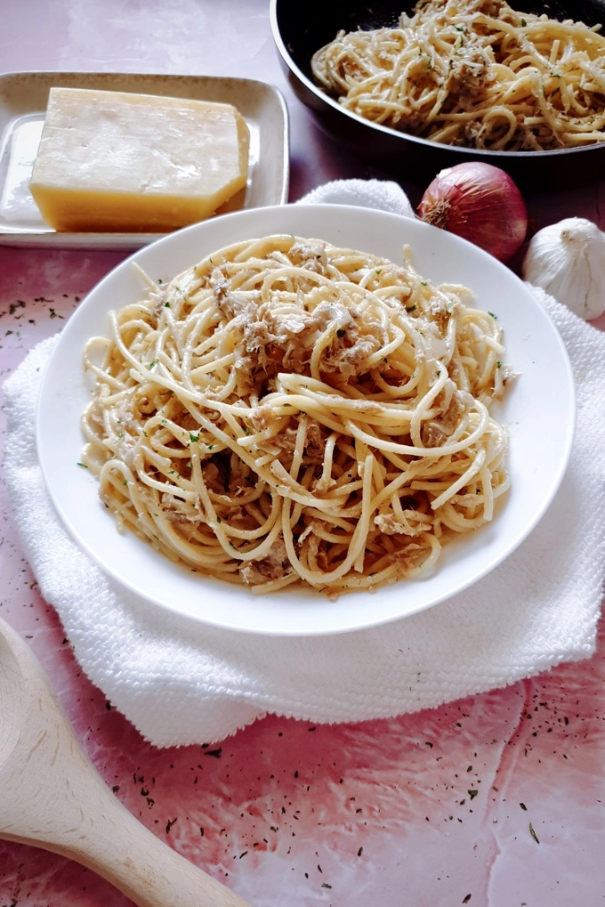 BEST and EASY Creamy Garlic Canned Tuna Pasta Recipe in a white plate and skillet, and a whole garlic, an onion, a Parmesan cheese block, a wooden ladle around the plate