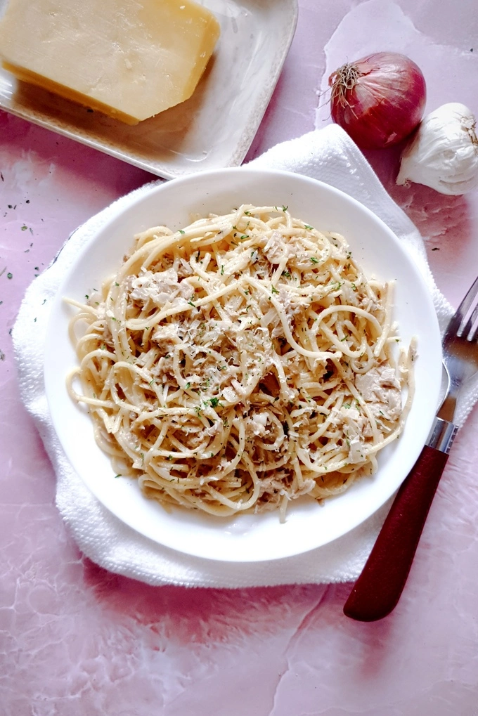 Creamy Garlic Canned Tuna Pasta Recipe served in a white plate over a white cloth, and a whole garlic, onion, a Parmesan cheese block and a fork next to the plate