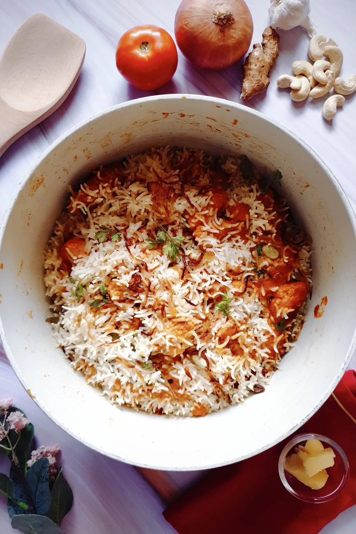 Butteriest Scrumptious Butter Chicken Biryani in a Dutch oven surrounded by a tomato, onion, ginger, garlic, cashew nuts, butter and a wooden ladle