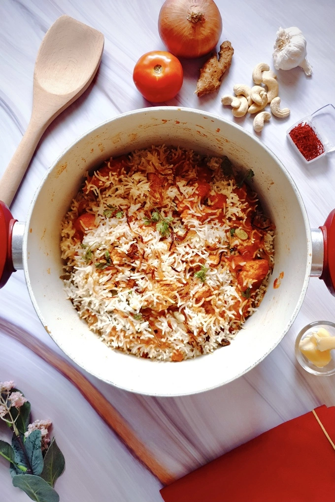 Butteriest Scrumptious Butter Chicken Biryani in a Dutch oven surrounded by ingredients like a tomato, onion, garlic, ginger, cashew nuts, small nowl of butter and saffron and a wooden ladle, red cloth and flower for decor
