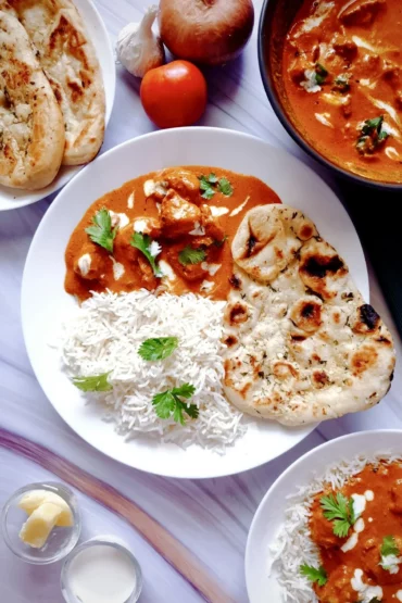 Dreamy Creamy One Pot Mouthwatering Butter Chicken Curry in a black bowl surrounded by a plate of steamed basmati rice, three butter garlic naans, butter and cream in two separate small bowls, a wooden ladle and some raw ingredients