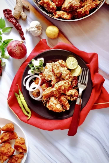 Easy Crispy Fried Indian Chicken Mixed with Tangy Sauce on a black plate placed on a red cloth and surrounded by a pan and plate with some cripsy chicken and a few whole ingredients like an onion, ginger, garlic, lemon, a sprig of curry leaves and two dried red chilies