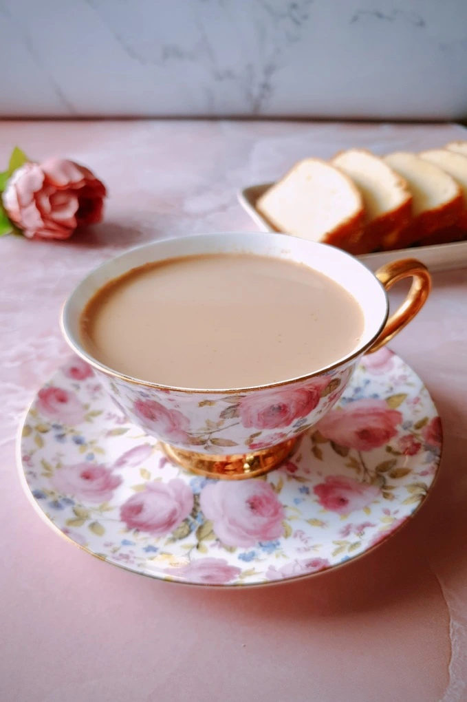 Homemade Japanese Royal Milk Tea in a fancy teacup over a plate and in the background a pink rose on the left for decor and on the right a plate of slices of butter cake