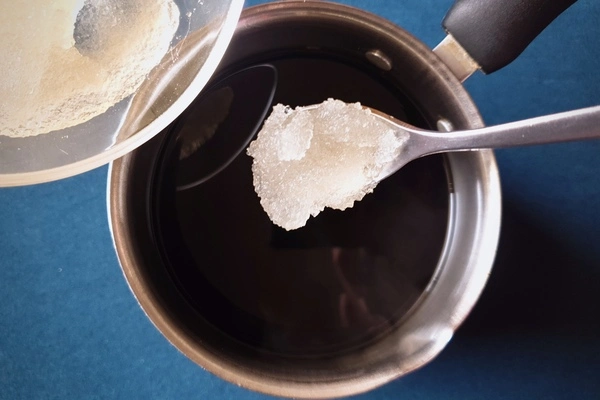 Top view of a saucepan with black coffee in it and a bowl of bloomed gelatin powder being poured from the side with a teaspoon