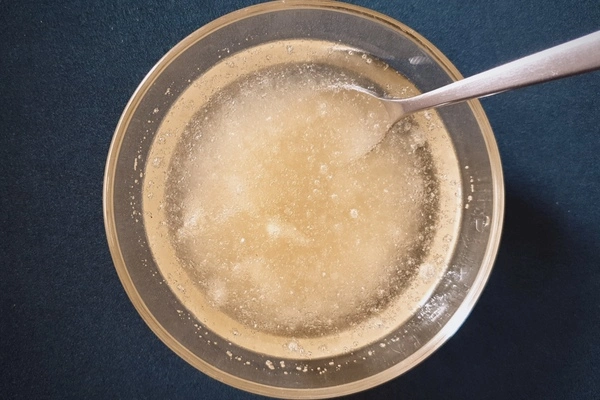 Top view of a glass bowl with water and gelatin powder in it being mixed with a teaspoon