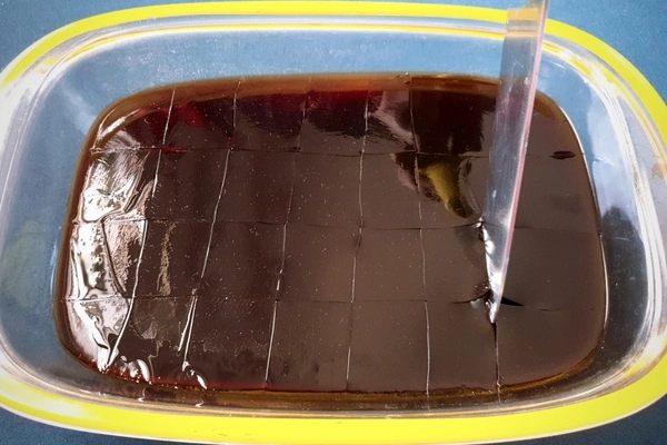 Coffee jelly in a glass tray being cut in horizontal and vertical lines into cubes with a knife