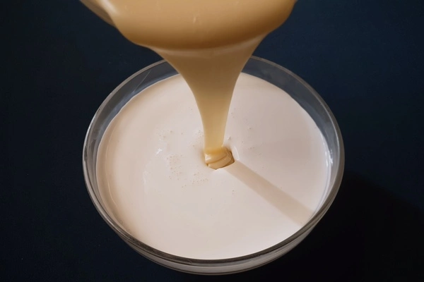 A glass bowl filled with heavy cream in it and another glass bowl right above it filled with sweetened condensed milk being poured in the heavy cream