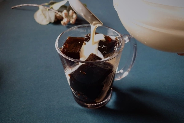 Cubes of coffee jello filled a glass mug with sweet condensed milk and cream mixture being poured in with a spoon