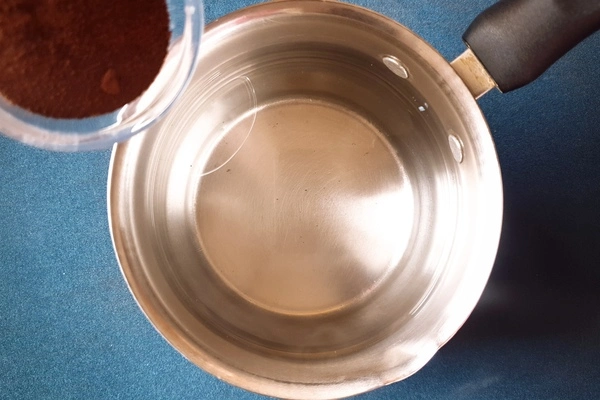 Top view of a saucepan with water in it and a small bowl of instant coffee powder being poured in