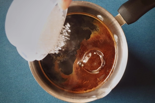 Top view of a saucepan with water and instant coffee powder in it and a small bowl of sugar being poured in
