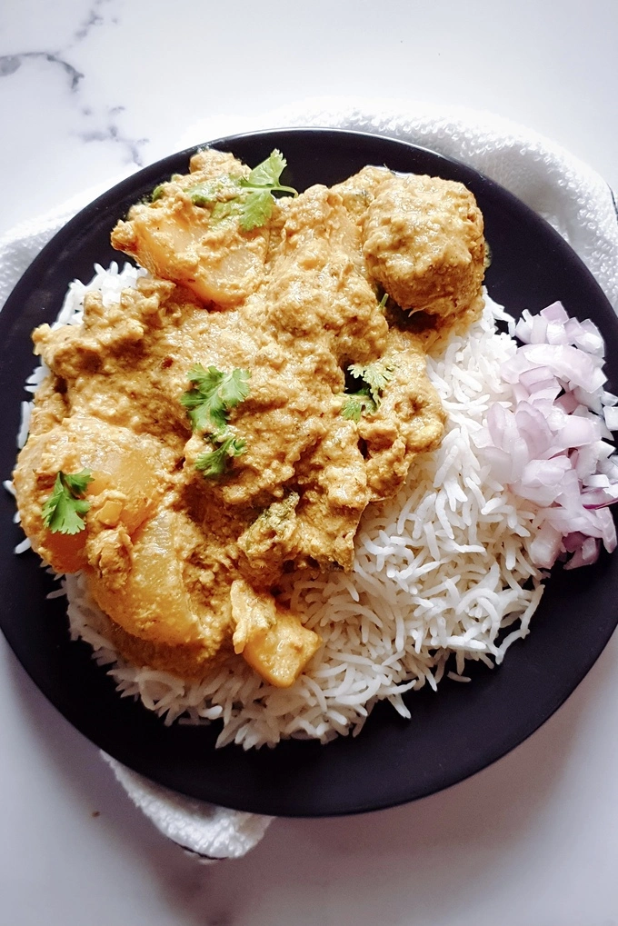 Rich and Aromatic Indian Chicken Korma Curry Cooked in Yogurt next to white basmati rice and chopped onions all served on a black plate over a white napkin
