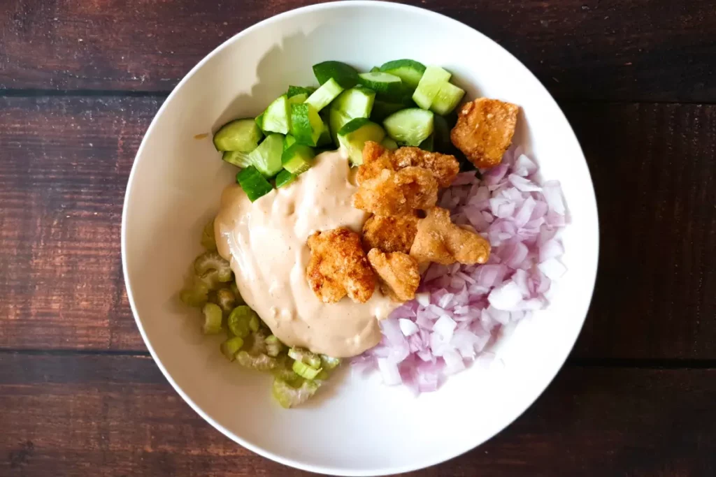 Chopped vegetables, salad dressing and fried pieces of chicken in a white mixing bowl.