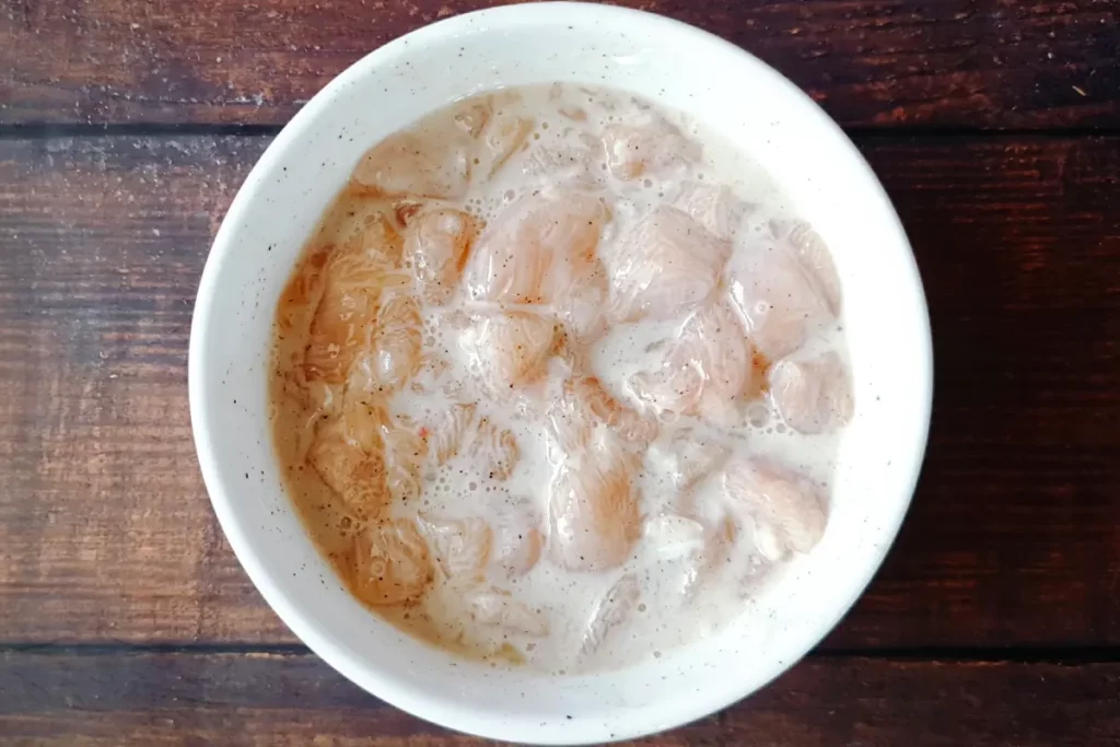 Small raw pieces of chicken in a white bowl along with other ingredients added in and mixed well to make fried chicken.